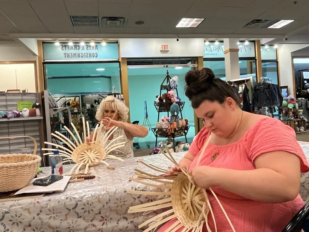 Two women seated at a table weaving spoked rattan basket bases, with mall storefronts behind them.