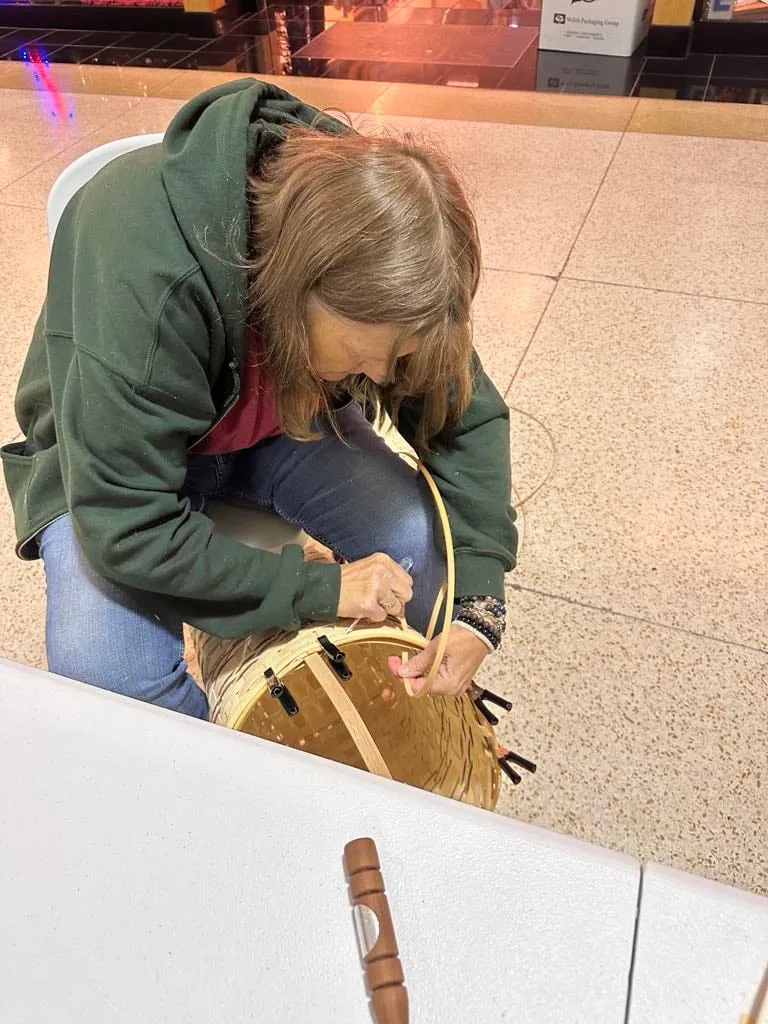 A woman in a green hoodie leaning over her work, finishing the rim of a basket.