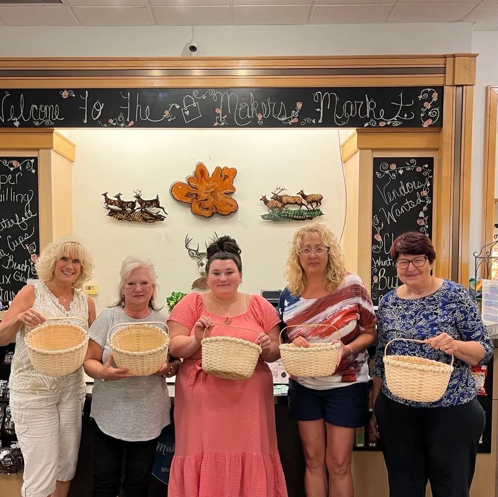 Five women lined up in front of the shop's chalkboard wall, each holding a finished woven basket they made in class.