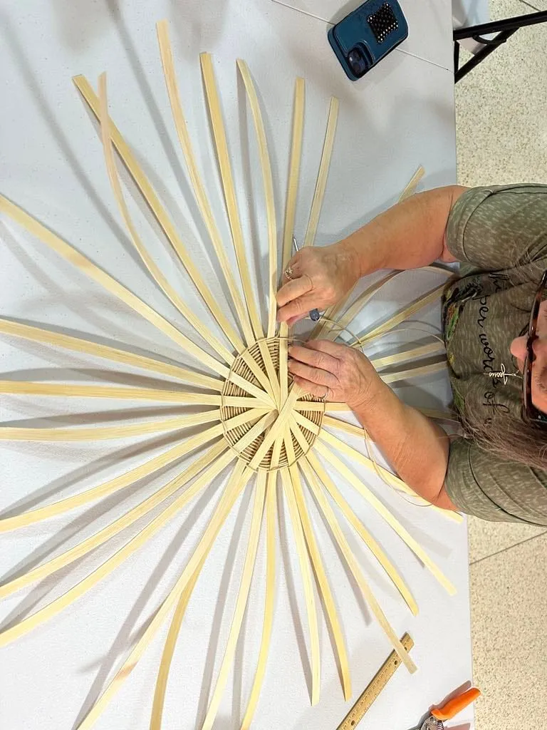 Top-down view of hands arranging a starburst of flat reed spokes into a woven base.