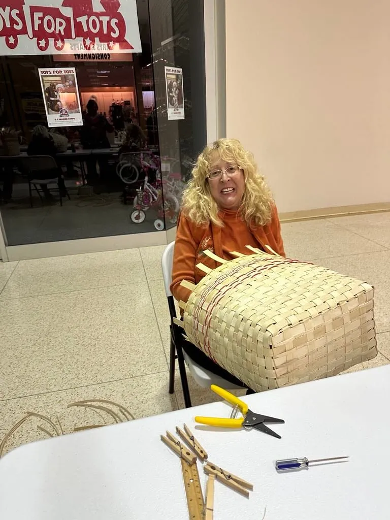 Woman seated behind a large flat woven piece with clothespins and pliers on the table.