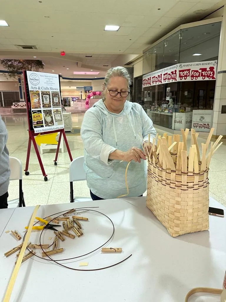 Woman standing at a work table weaving a tall basket held together with clothespin clamps.