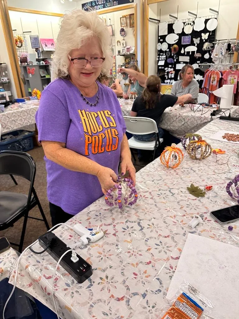 Woman in a purple 'Hocus Pocus' t-shirt assembling a beaded pumpkin at a floral-cloth table.