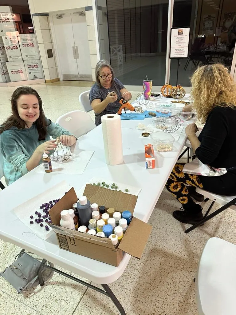 Several women around a long white table with a supply box of paint and bead bottles in the foreground.