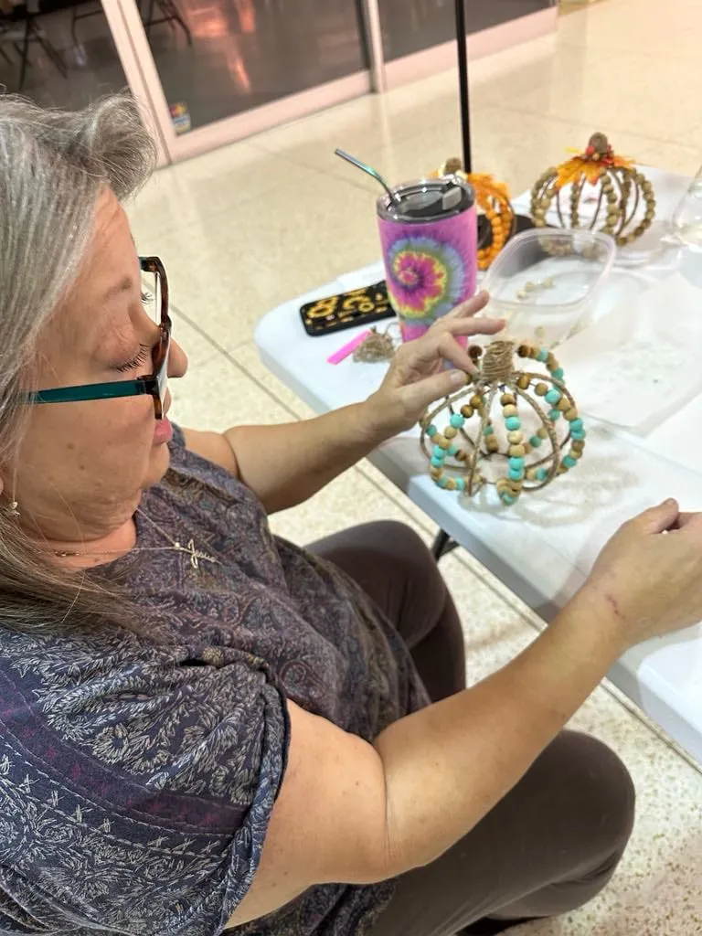 Woman with glasses working on a teal-and-white beaded pumpkin, a finished gold pumpkin beside her.