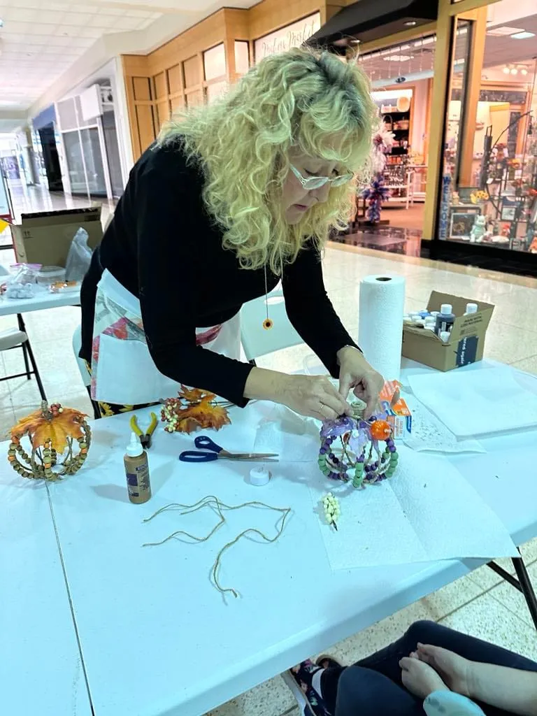Woman leaning over the table assembling a multicolor beaded pumpkin, wire and scissors beside her.