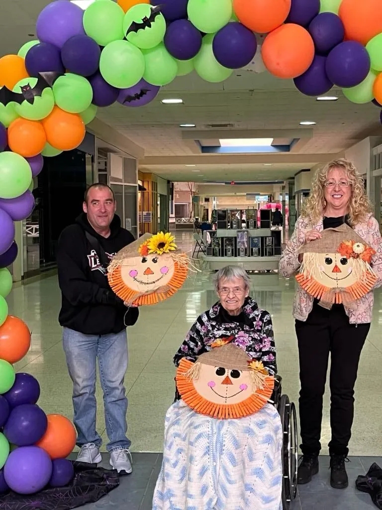 Four participants holding finished burlap scarecrow face crafts under a purple, green and orange Halloween balloon arch.