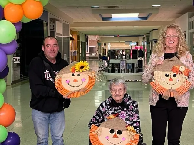 Four people holding up the finished burlap scarecrow faces they made, standing under a purple, green and orange balloon arch.