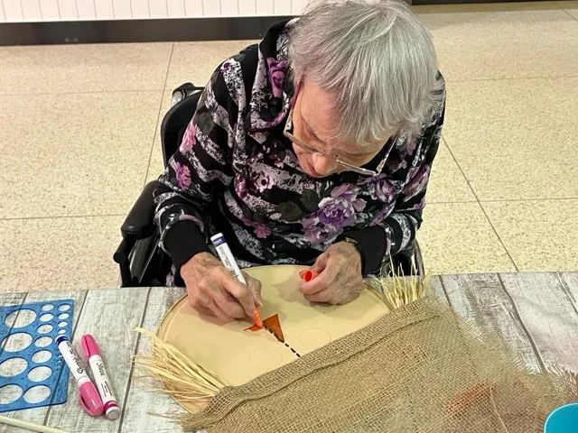 A woman at the craft table drawing a face onto a burlap scarecrow panel, with markers and raffia laid out beside her.