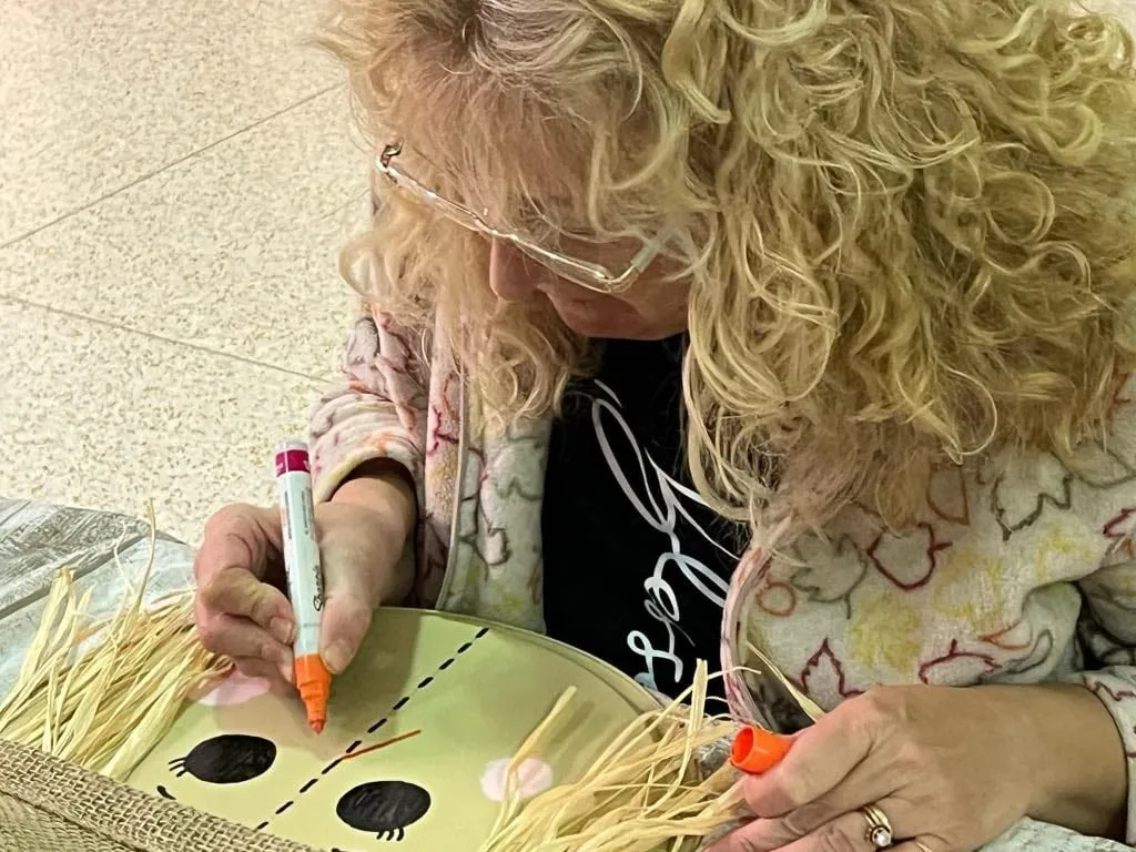 Woman drawing large black eyes onto a green-painted scarecrow face with raffia hair.