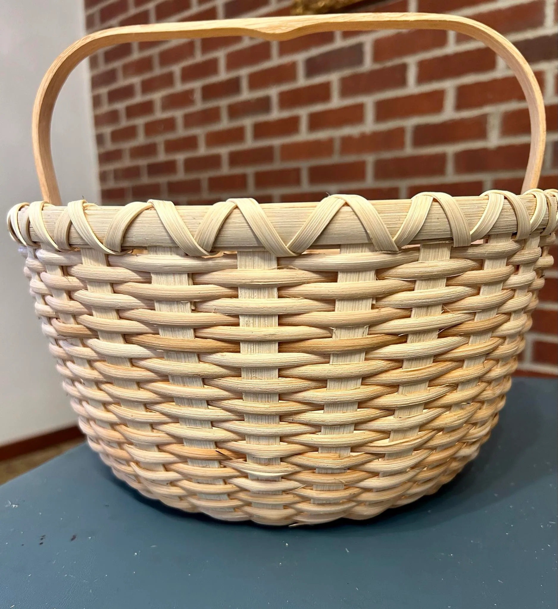 Finished round natural-reed basket with a bent wood swing handle and a chevron accent band, against a brick wall.