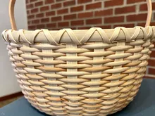 Finished round natural-reed basket with a bent wood swing handle and a chevron accent band, photographed against a brick wall.