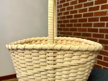 Finished rectangular market basket with a tall arched handle and plain woven body, resting on a slate surface against a brick wall.