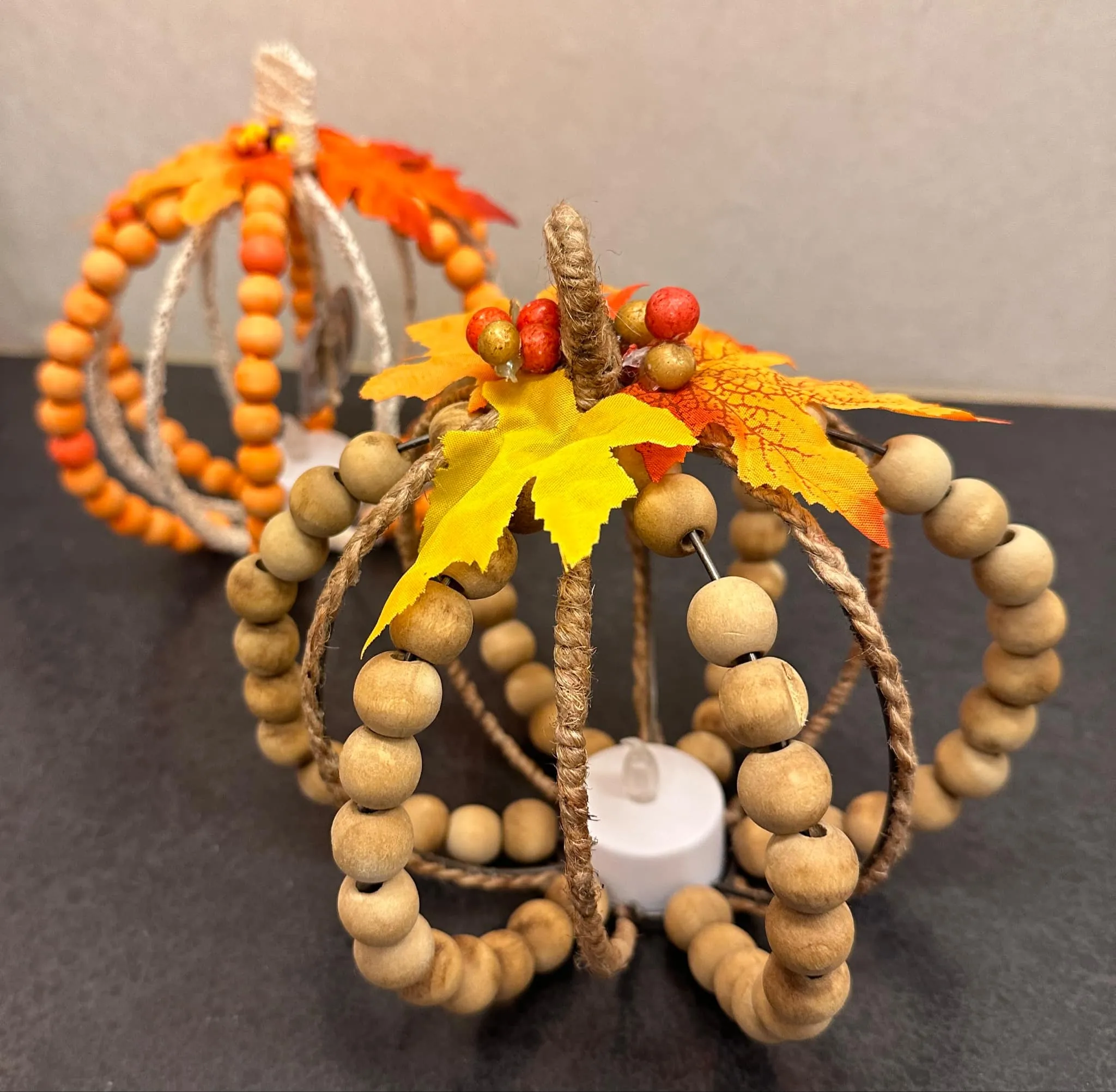 Two finished pumpkins made from natural wood beads and twine, topped with orange and yellow silk maple leaves and berry sprigs, lit from inside by a small LED tealight.