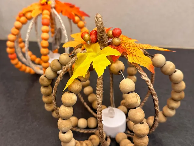Two finished pumpkins made from natural wood beads and twine, topped with silk maple leaves and lit from inside by a small tealight.